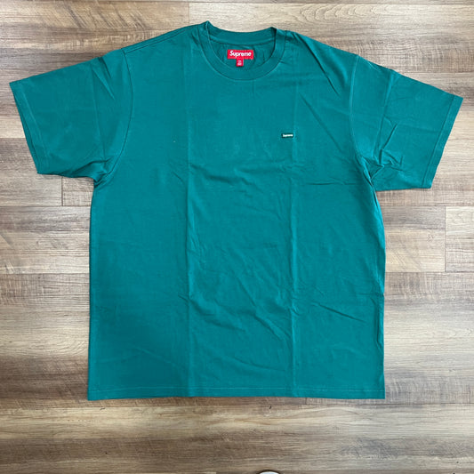 A Supreme Small Box Tee (FW23) Light Pine, preowned with a small rectangular black tag on the front and a red neck tag with white text, is laid flat on a wooden floor, displaying its excellent condition.