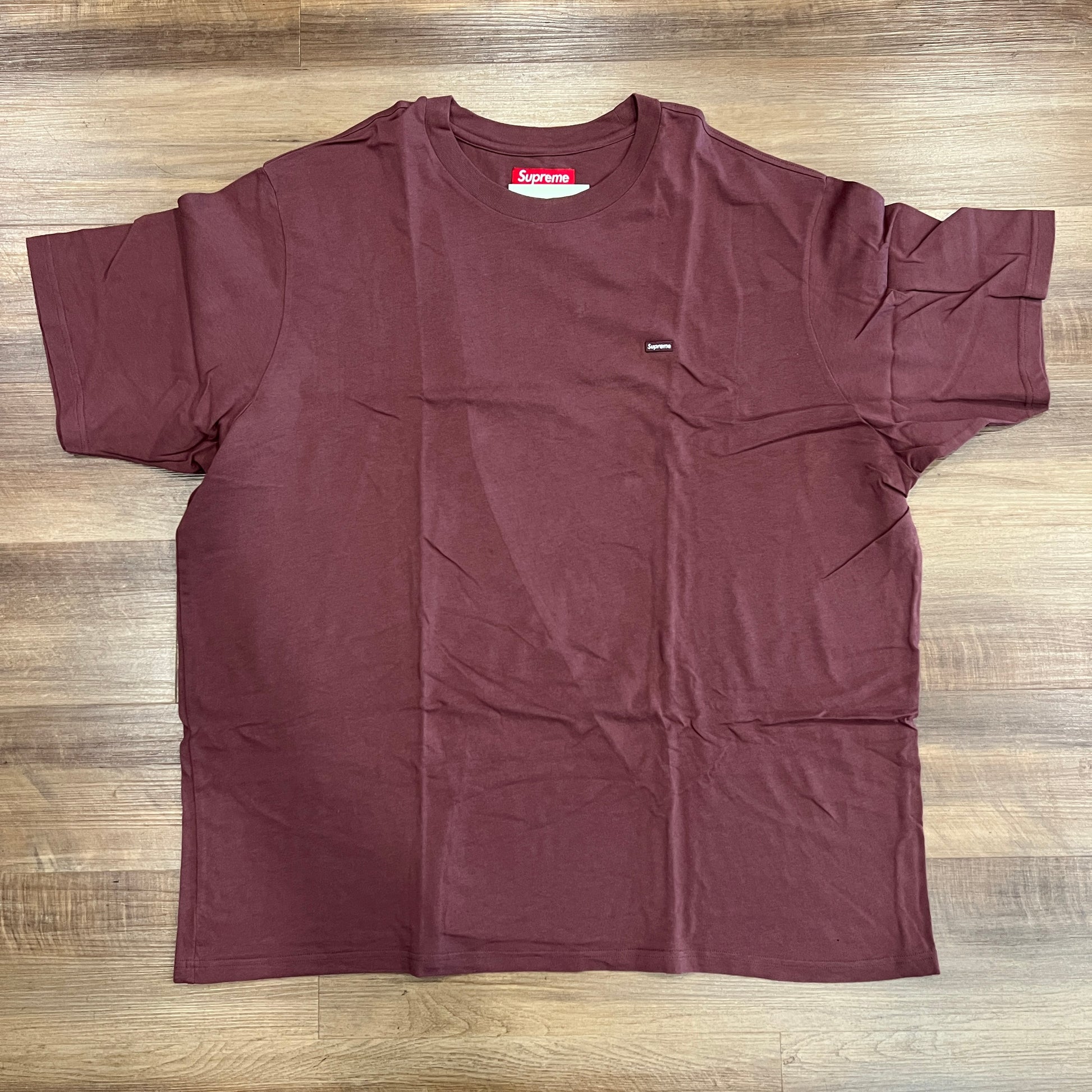 A preowned Supreme Small Box Tee (FW23) in washed plum is laid out on a wooden floor, showcasing its small chest logo and a red collar tag.