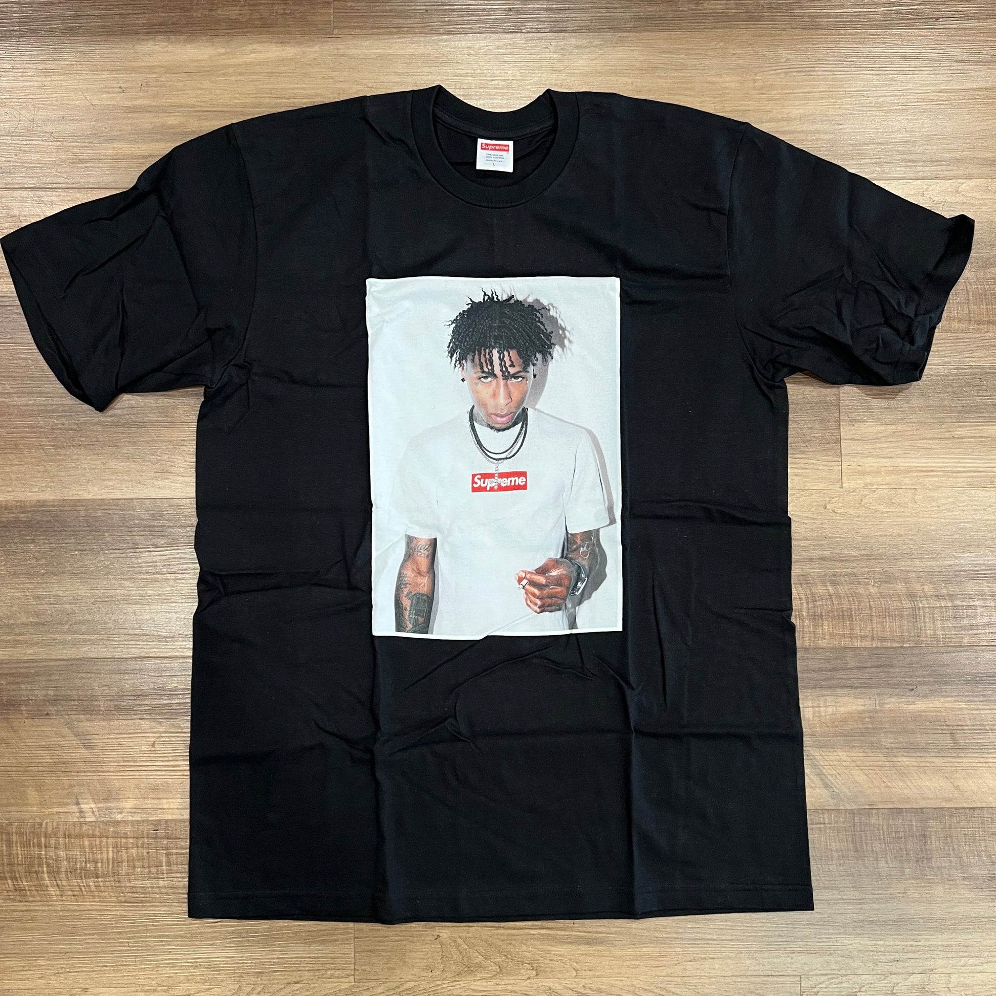 In new condition, the Supreme NBA Youngboy Tee Black features a graphic of someone in a white tee with Supremes red logo, holding a lighter and facing forward. The shirt is neatly laid flat on a wooden floor.