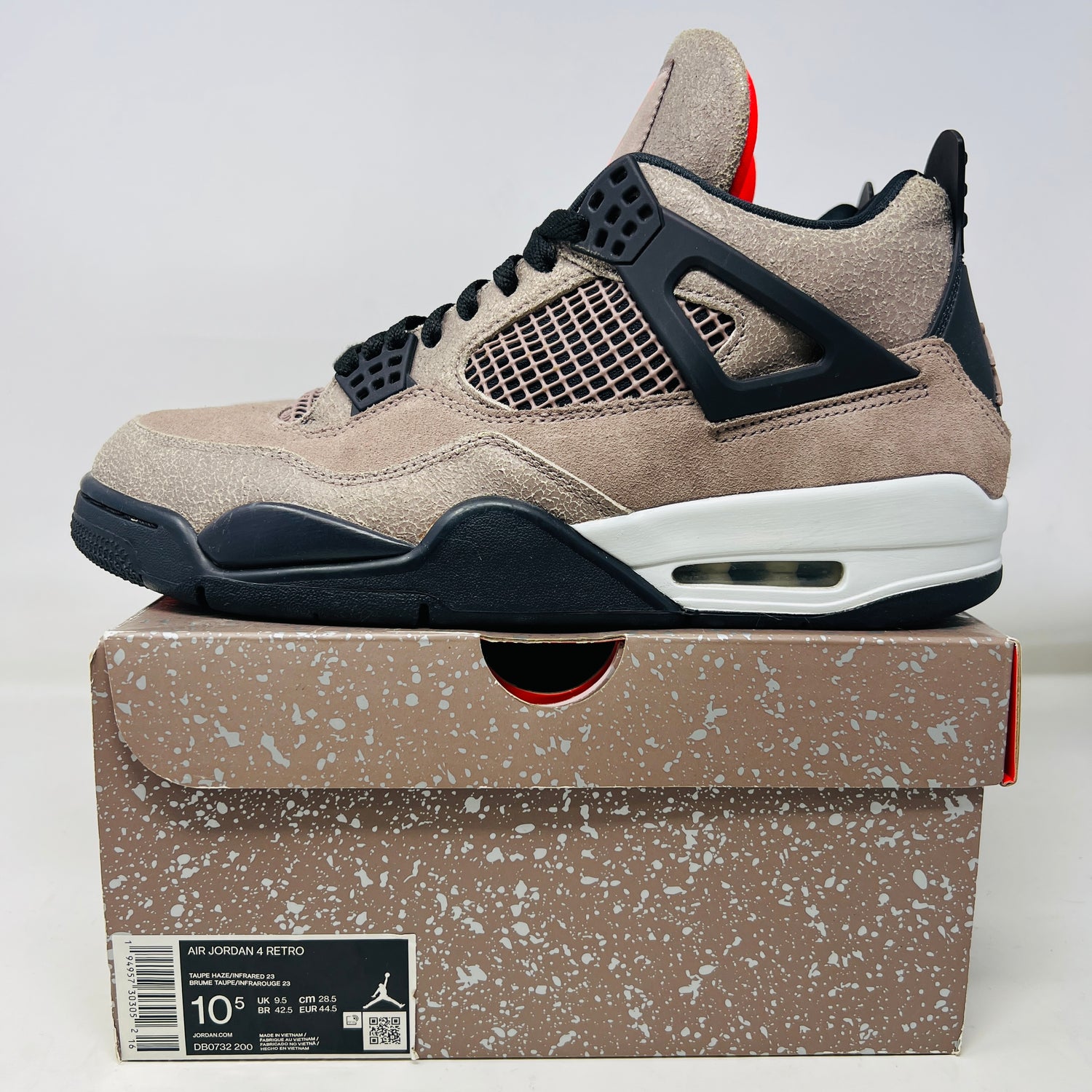 A tan Jordan 4 Retro Taupe Haze sneaker sits on a matching speckled box, featuring clean uppers with black netting, laces, and accents. The look is finished with a white and black sole. Box label details include shoe size.