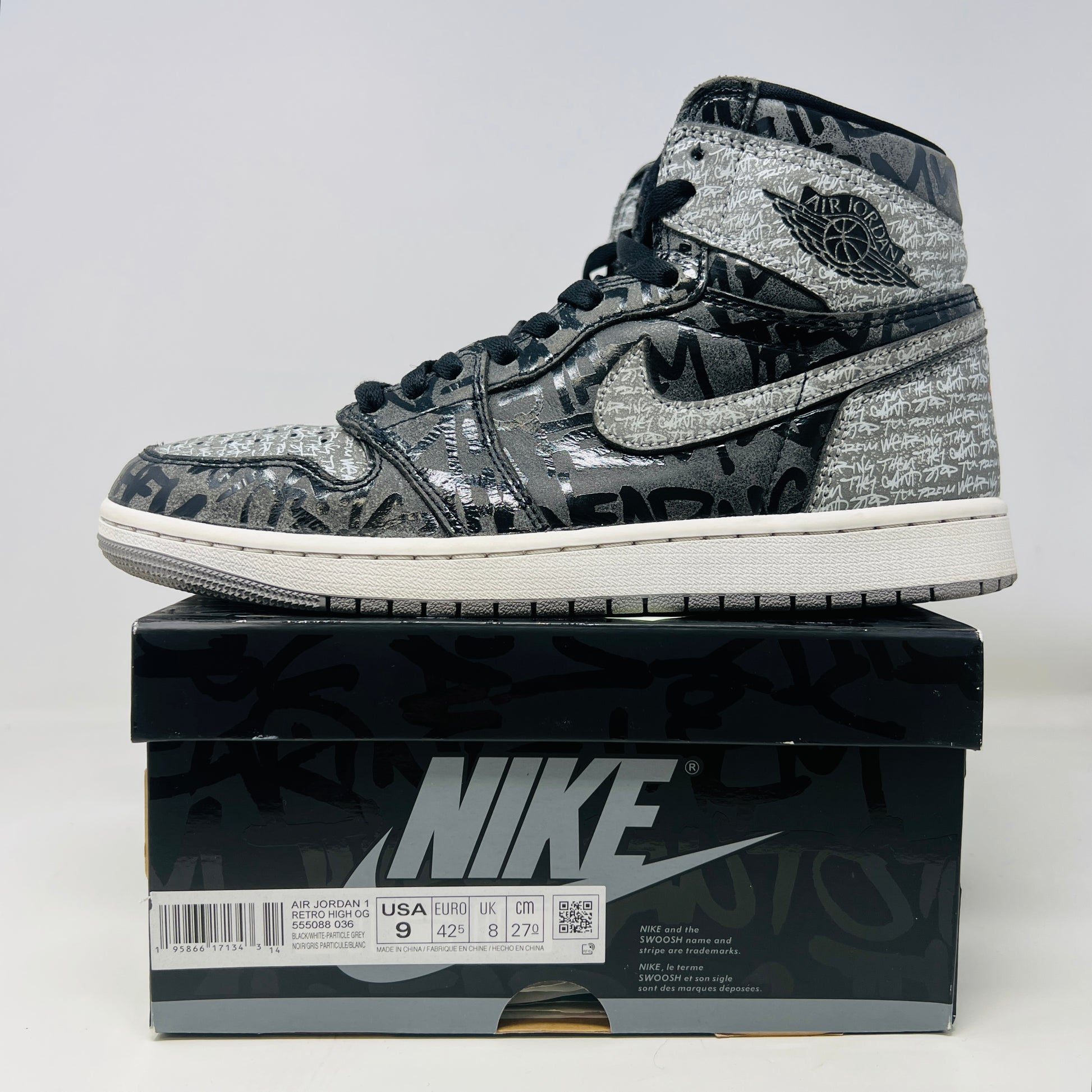 A gray and black Jordan 1 Retro High OG Rebellionaire sneaker with a graffiti-style design sits on a black Nike shoebox featuring the Nike logo and size details. Slight heel drag indicates its journey.