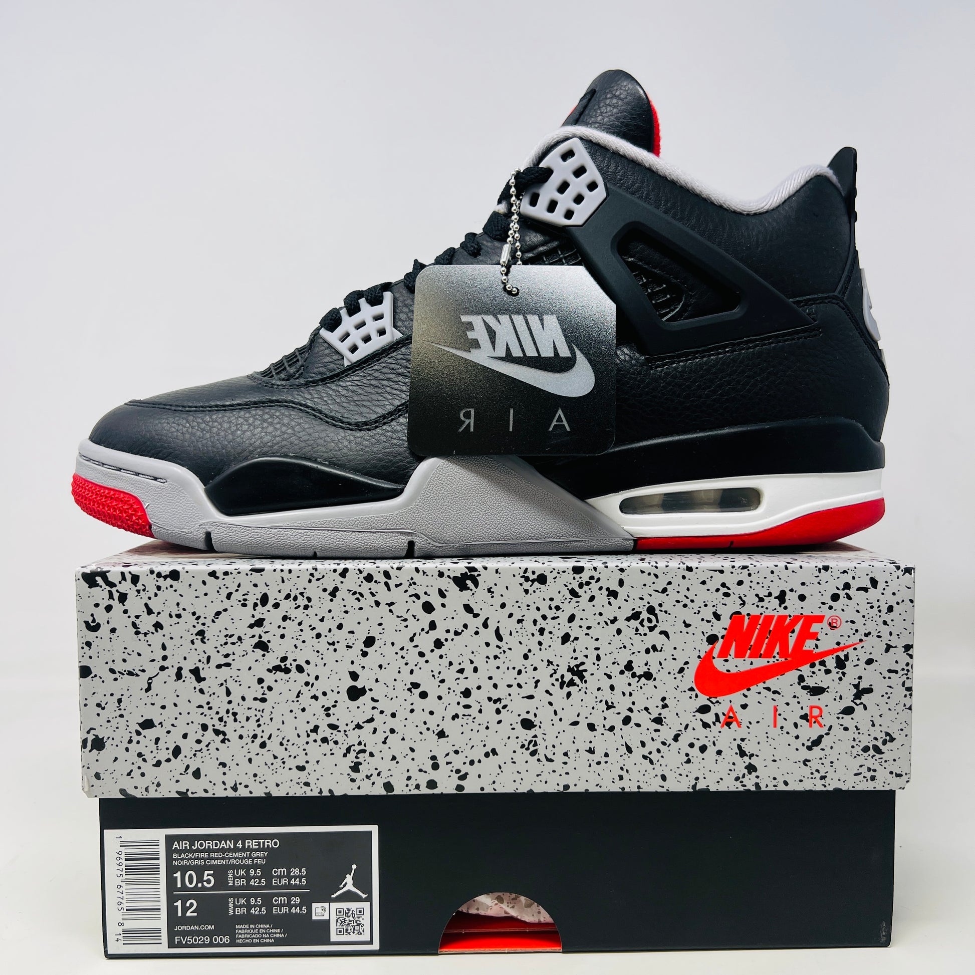 A pair of Jordan 4 Retro Bred Reimagined sneakers, featuring black with red and white accents, is displayed atop a matching speckled shoebox. The shoe has a Nike Air tag attached, and the pristine box showcases all accessories along with size and product details.