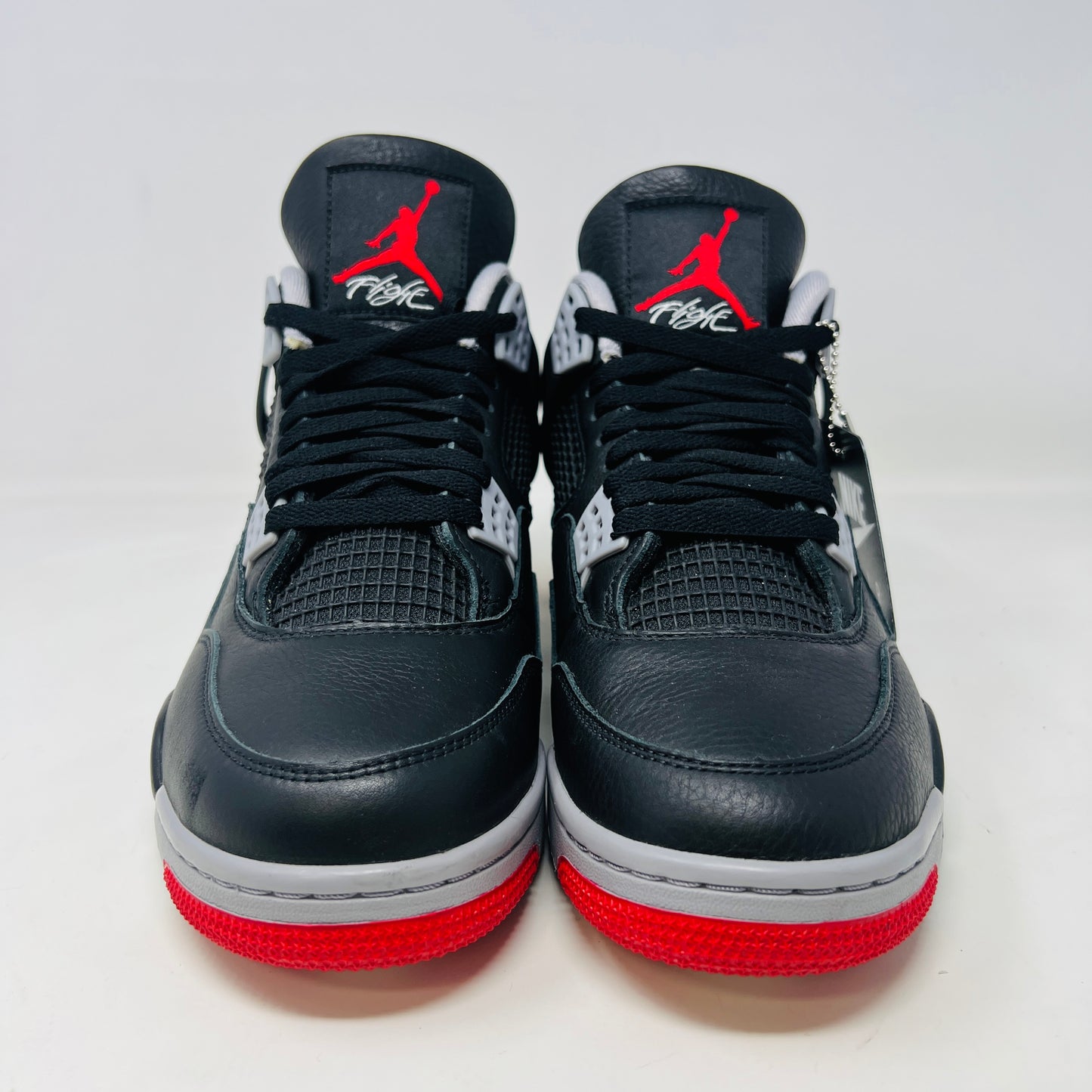 A pair of Jordan 4 Retro Bred Reimagined sneakers, featuring black with red and white accents, is displayed atop a matching speckled shoebox. The shoe has a Nike Air tag attached, and the pristine box showcases all accessories along with size and product details.