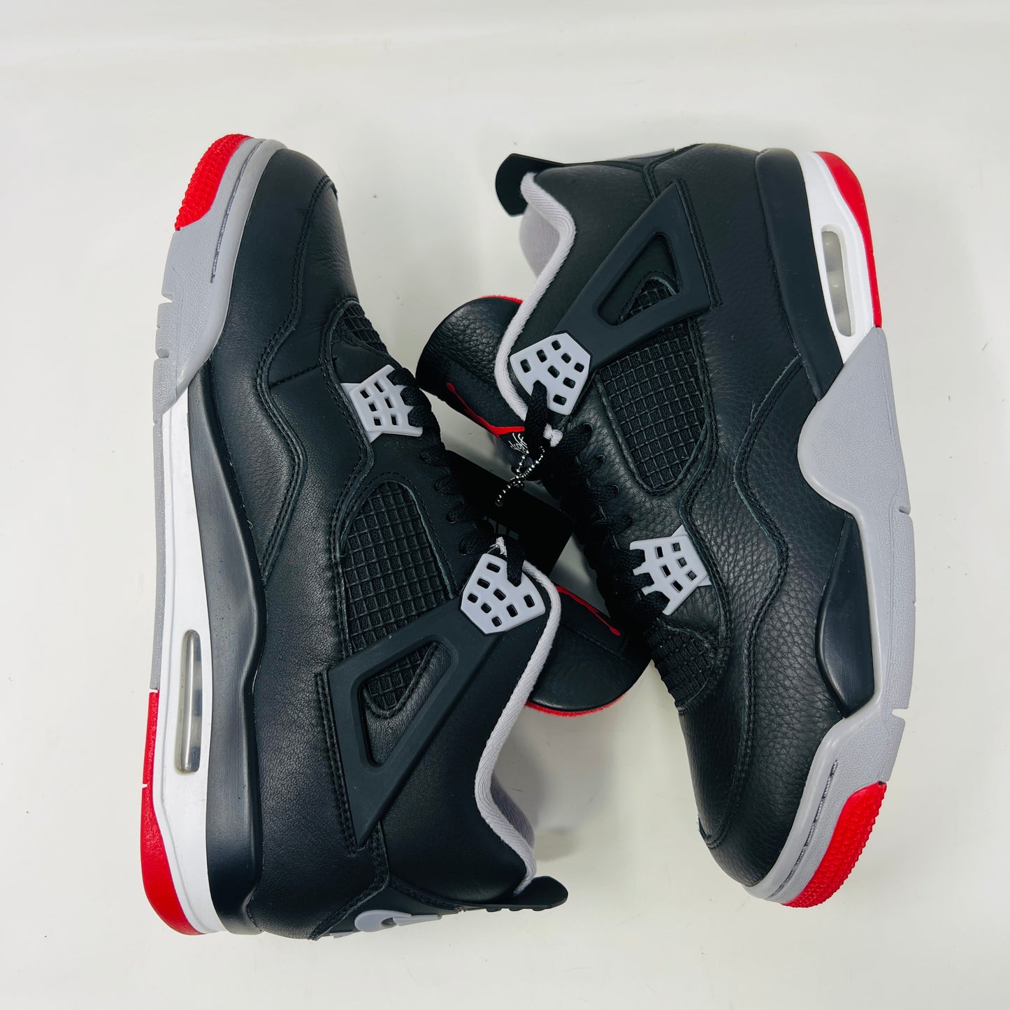 A pair of Jordan 4 Retro Bred Reimagined sneakers, featuring black with red and white accents, is displayed atop a matching speckled shoebox. The shoe has a Nike Air tag attached, and the pristine box showcases all accessories along with size and product details.