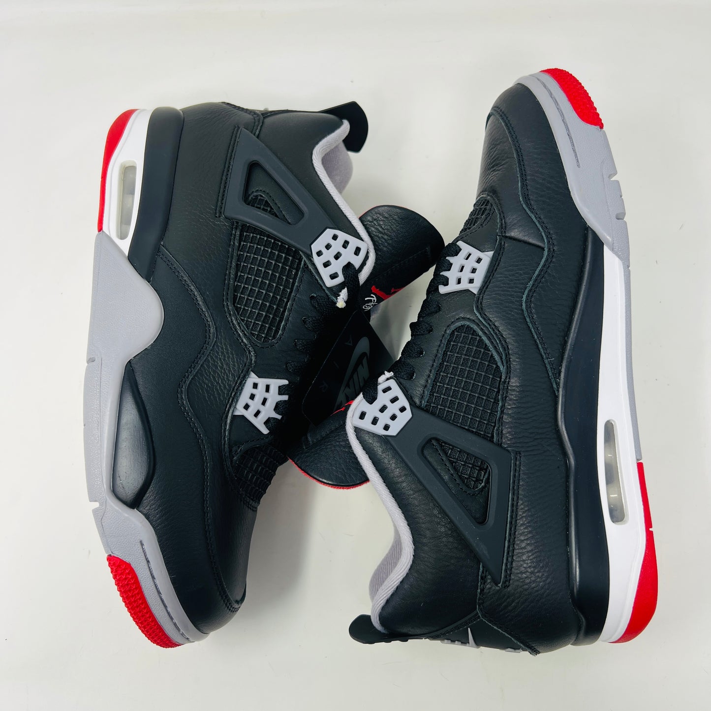 A pair of Jordan 4 Retro Bred Reimagined sneakers, featuring black with red and white accents, is displayed atop a matching speckled shoebox. The shoe has a Nike Air tag attached, and the pristine box showcases all accessories along with size and product details.