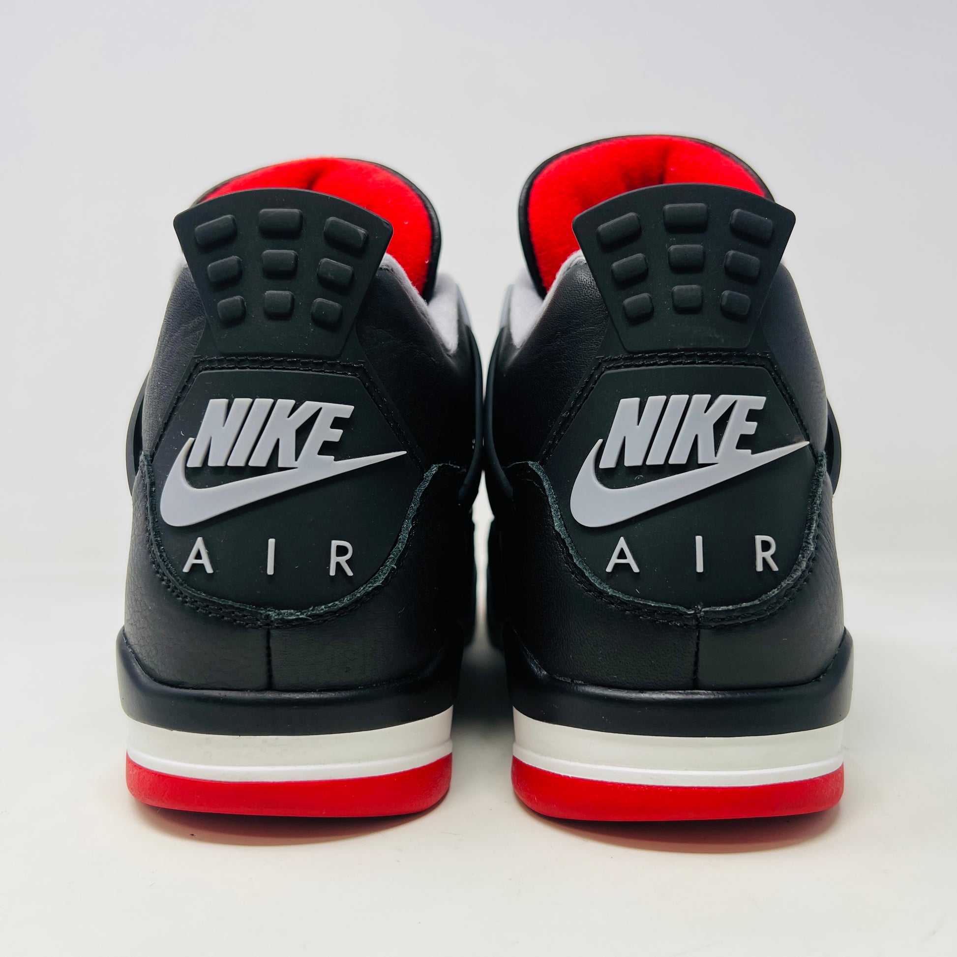 A pair of Jordan 4 Retro Bred Reimagined sneakers, featuring black with red and white accents, is displayed atop a matching speckled shoebox. The shoe has a Nike Air tag attached, and the pristine box showcases all accessories along with size and product details.