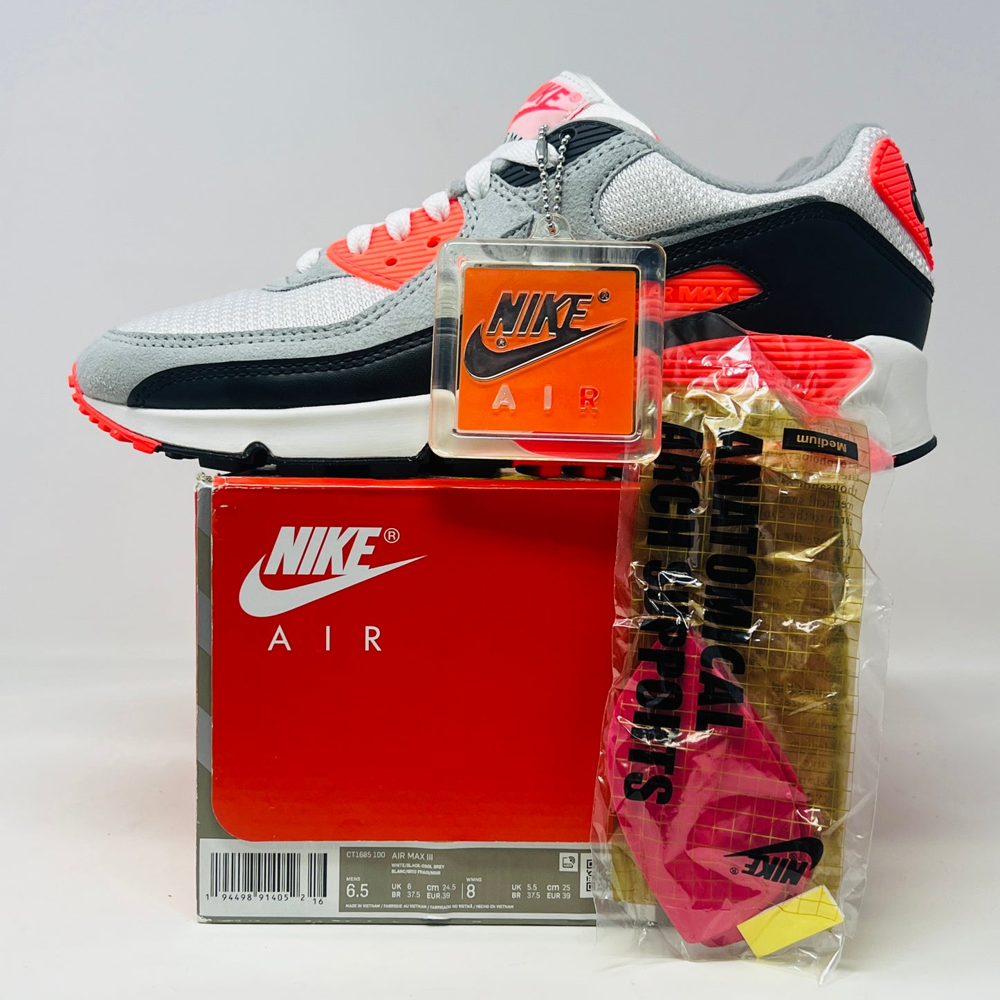 The Nike Air Max 90 Infrared (2020) sneaker in grey, black, and red sits on a red Nike box. Nearby are Anatomically Arched Orthotics insoles in a clear package. The shoe showcases clean uppers and an attached hang tag for a sleek look.