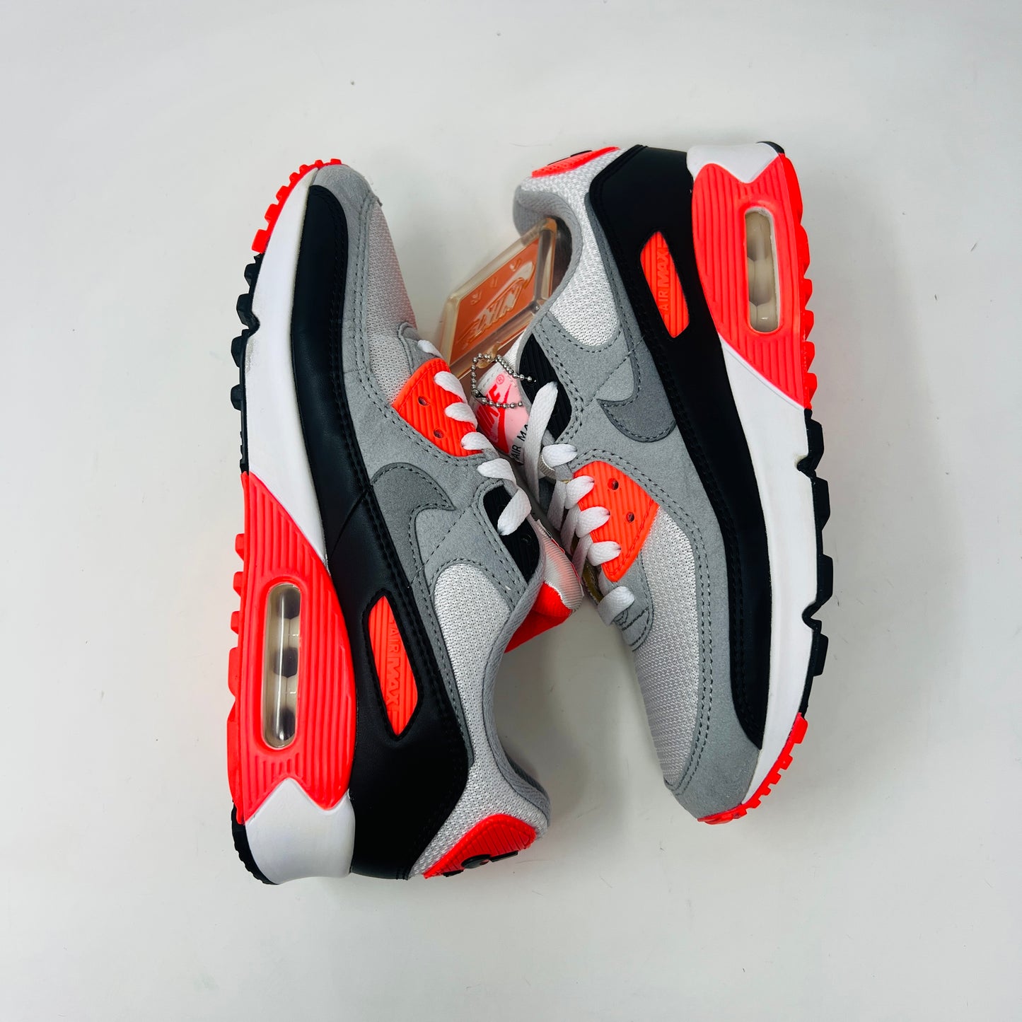 The Nike Air Max 90 Infrared (2020) sneaker in grey, black, and red sits on a red Nike box. Nearby are Anatomically Arched Orthotics insoles in a clear package. The shoe showcases clean uppers and an attached hang tag for a sleek look.