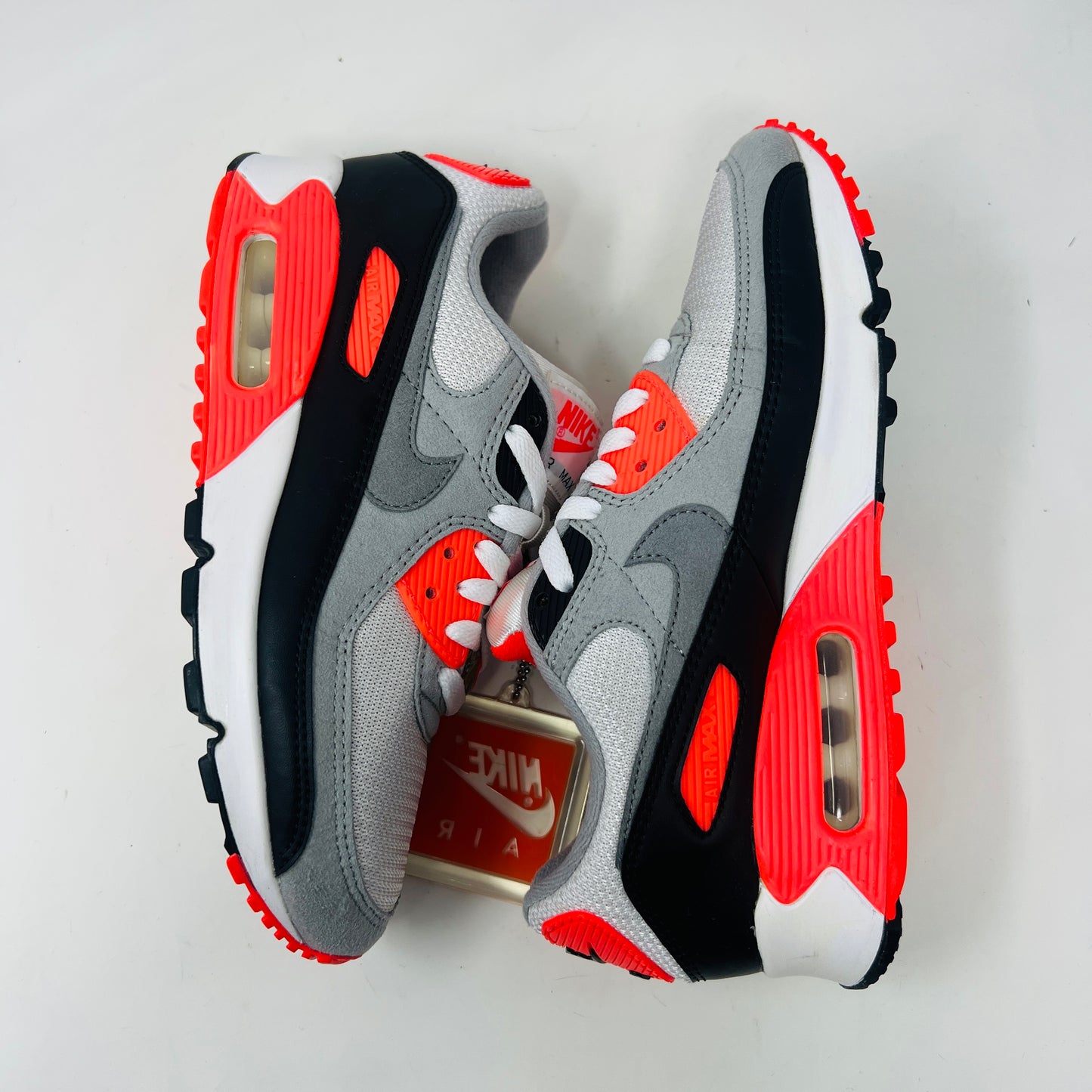 The Nike Air Max 90 Infrared (2020) sneaker in grey, black, and red sits on a red Nike box. Nearby are Anatomically Arched Orthotics insoles in a clear package. The shoe showcases clean uppers and an attached hang tag for a sleek look.