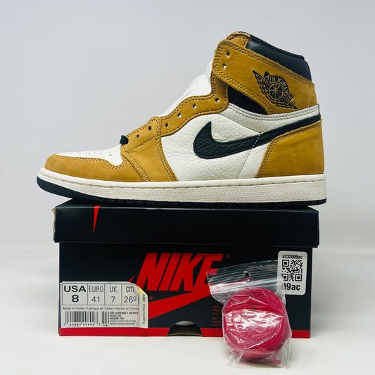 The Jordan 1 Retro High Rookie of the Year sneaker by Jordan, in white and gold with black detailing and swoosh, rests on a black box. In front, a bag of extra red laces accompanies the box showing size and branding information.