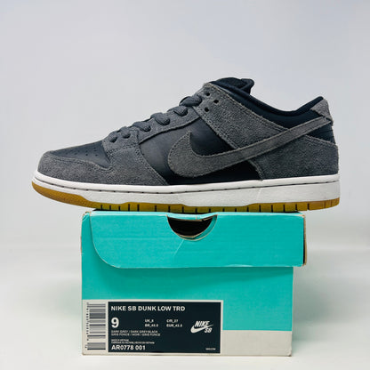 Displayed on a turquoise box, the 2018 Nike SB Dunk Low Dark Grey Black Gum shoe features gray suede uppers, a black Swoosh, and a gum sole. The slightly opened lid reveals product details inside.