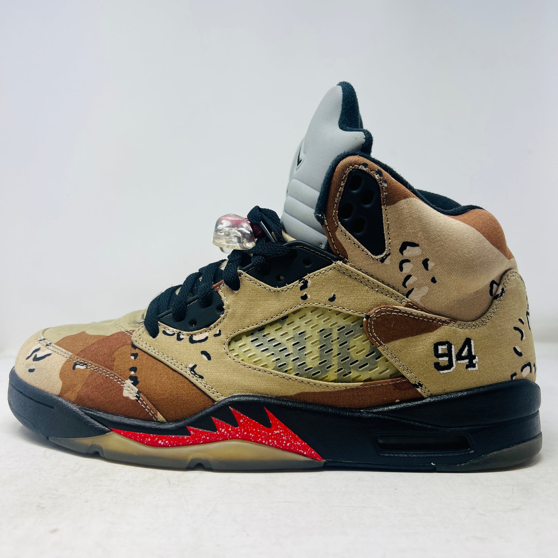 The Jordan 5 Retro Supreme Desert Camo is a high-top sneaker with tan, brown, and black camouflage. It features a clean upper, black laces with locks, a mesh side panel, gray tongue, red midsole accents, and 94 on the heel for an authentic touch.