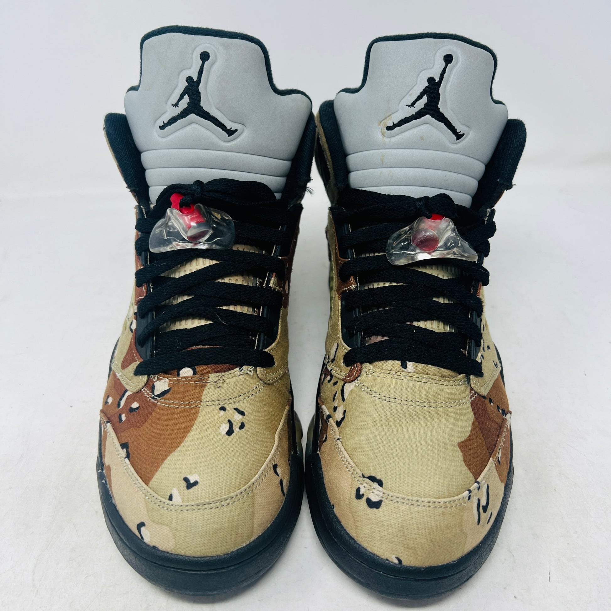 The Jordan 5 Retro Supreme Desert Camo is a high-top sneaker with tan, brown, and black camouflage. It features a clean upper, black laces with locks, a mesh side panel, gray tongue, red midsole accents, and 94 on the heel for an authentic touch.