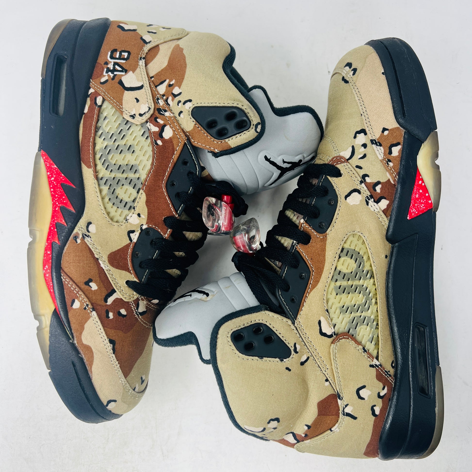 The Jordan 5 Retro Supreme Desert Camo is a high-top sneaker with tan, brown, and black camouflage. It features a clean upper, black laces with locks, a mesh side panel, gray tongue, red midsole accents, and 94 on the heel for an authentic touch.