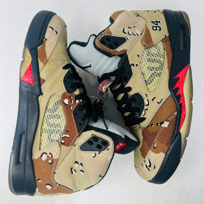 The Jordan 5 Retro Supreme Desert Camo is a high-top sneaker with tan, brown, and black camouflage. It features a clean upper, black laces with locks, a mesh side panel, gray tongue, red midsole accents, and 94 on the heel for an authentic touch.