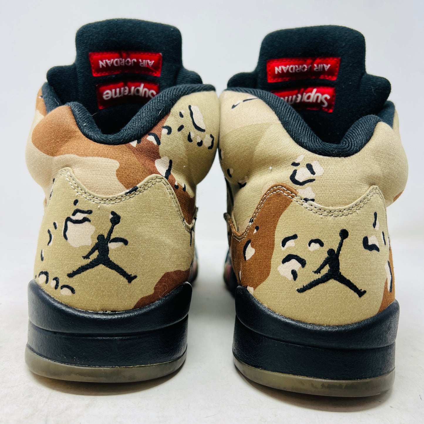 The Jordan 5 Retro Supreme Desert Camo is a high-top sneaker with tan, brown, and black camouflage. It features a clean upper, black laces with locks, a mesh side panel, gray tongue, red midsole accents, and 94 on the heel for an authentic touch.