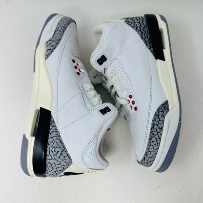 The Jordan 3 Retro White Cement Reimagined (GS) sneakers by Jordan boast a white leather upper with red accents, gray elephant print detailing, and a black heel and midsole, displayed on a white surface.