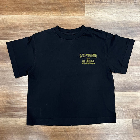 A Holy Ground black cropped Holy Rumble Tee, offered in sizes XS-XL, lies on a wooden floor. Bright yellow text on the left chest promotes The Big Cheese sporting event with captivating phrases like El Hombres.