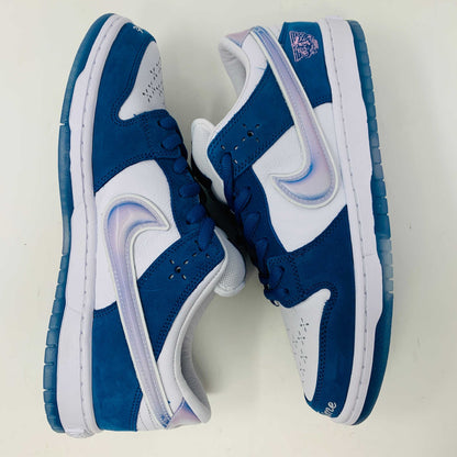 The Nike SB Dunk Low Born X Raised One Block At A Time sneaker, in blue and white with a metallic swoosh and embroidered heel details, sits on a Nike shoebox. It includes three pairs of shoelaces—two white and one pink—in front of the 2023 edition box.