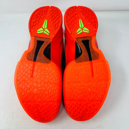 The image displays the Nike Kobe 6 Protro Reverse Grinch athletic shoes, featuring bright orange soles with a black section and a neon green logo near the heel. The hexagonal-patterned texture is shown as the shoes are placed sole-up on a white surface.