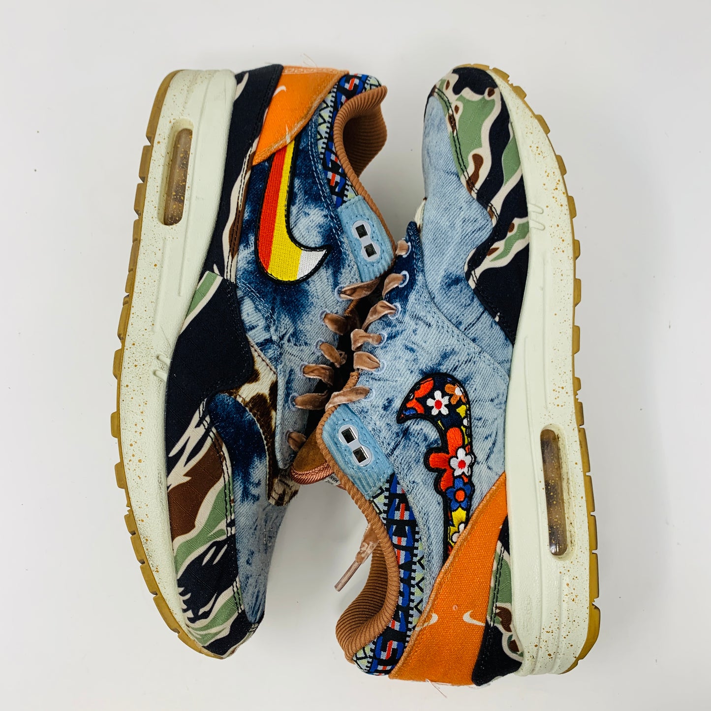 The Nike Air Max 1 SP Concepts Heavy sneakers feature a vibrant mixed pattern of camouflage, denim, and abstract shapes with a yellow swoosh, brown laces, and white soles with a speckled effect.