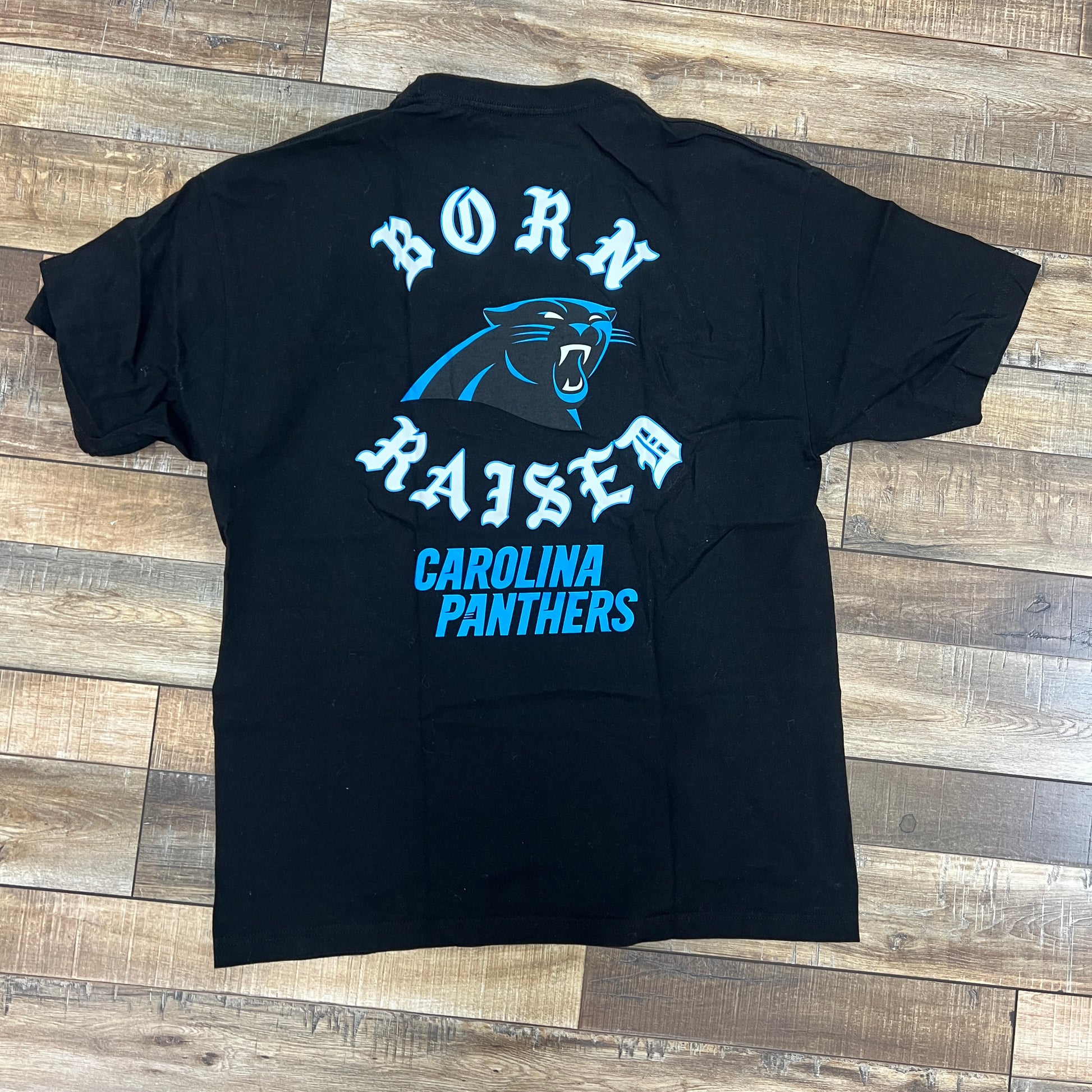 A preowned Born x Raised Carolina Panthers Tee in black is displayed on a wooden floor, showcasing the Born Raised logo in white encircling an authentic roaring panther. The words Carolina Panthers appear in blue beneath.