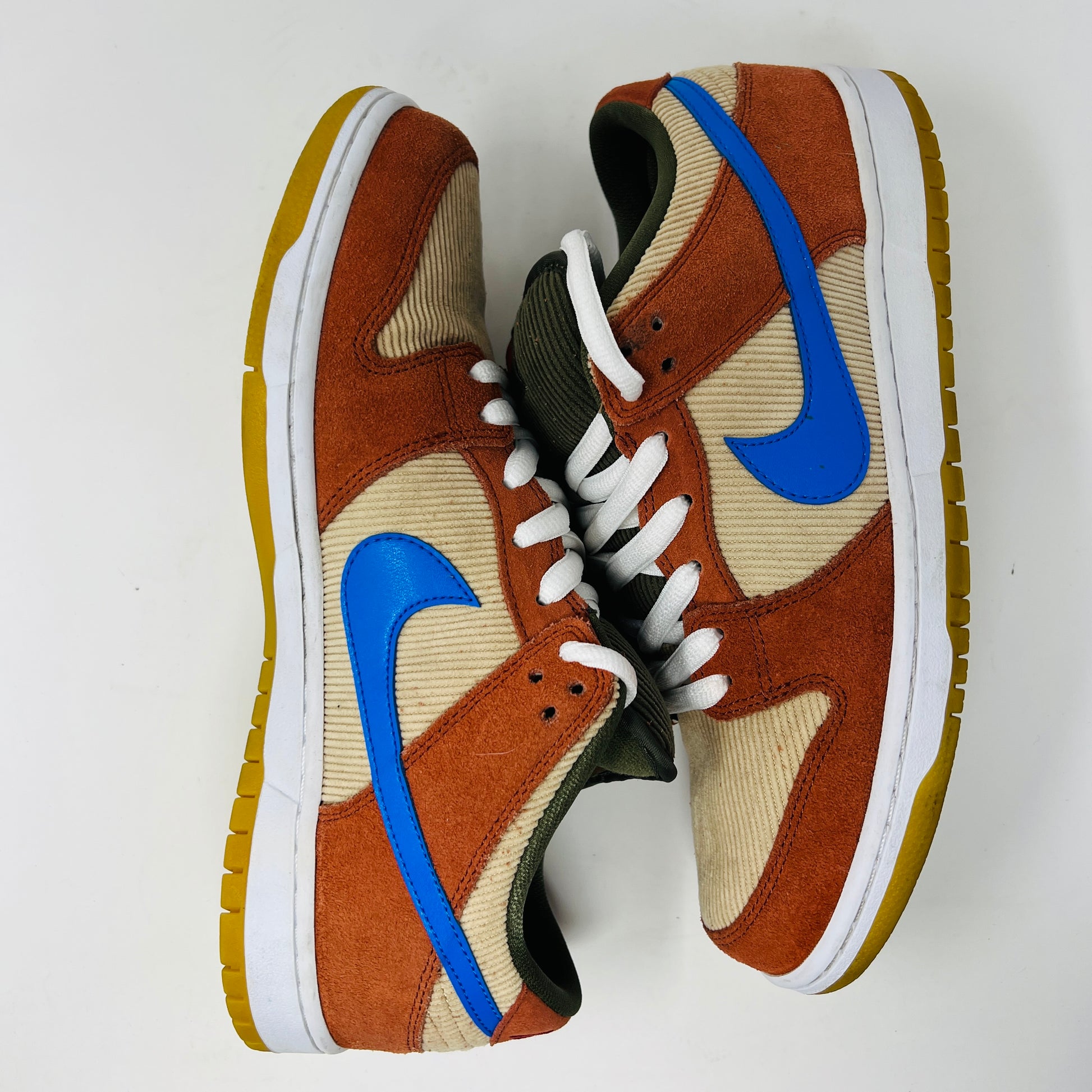 The Nike SB Dunk Low Corduroy Dusty Peach sneakers, by Nike, feature rust-orange and beige panels with bright blue swooshes, olive laces, yellow gum soles, and a textured fabric finish. They are displayed side by side on a white surface.