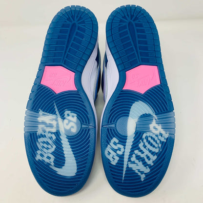 The Nike SB Dunk Low Born X Raised One Block At A Time sneaker, in blue and white with a metallic swoosh and embroidered heel details, sits on a Nike shoebox. It includes three pairs of shoelaces—two white and one pink—in front of the 2023 edition box.