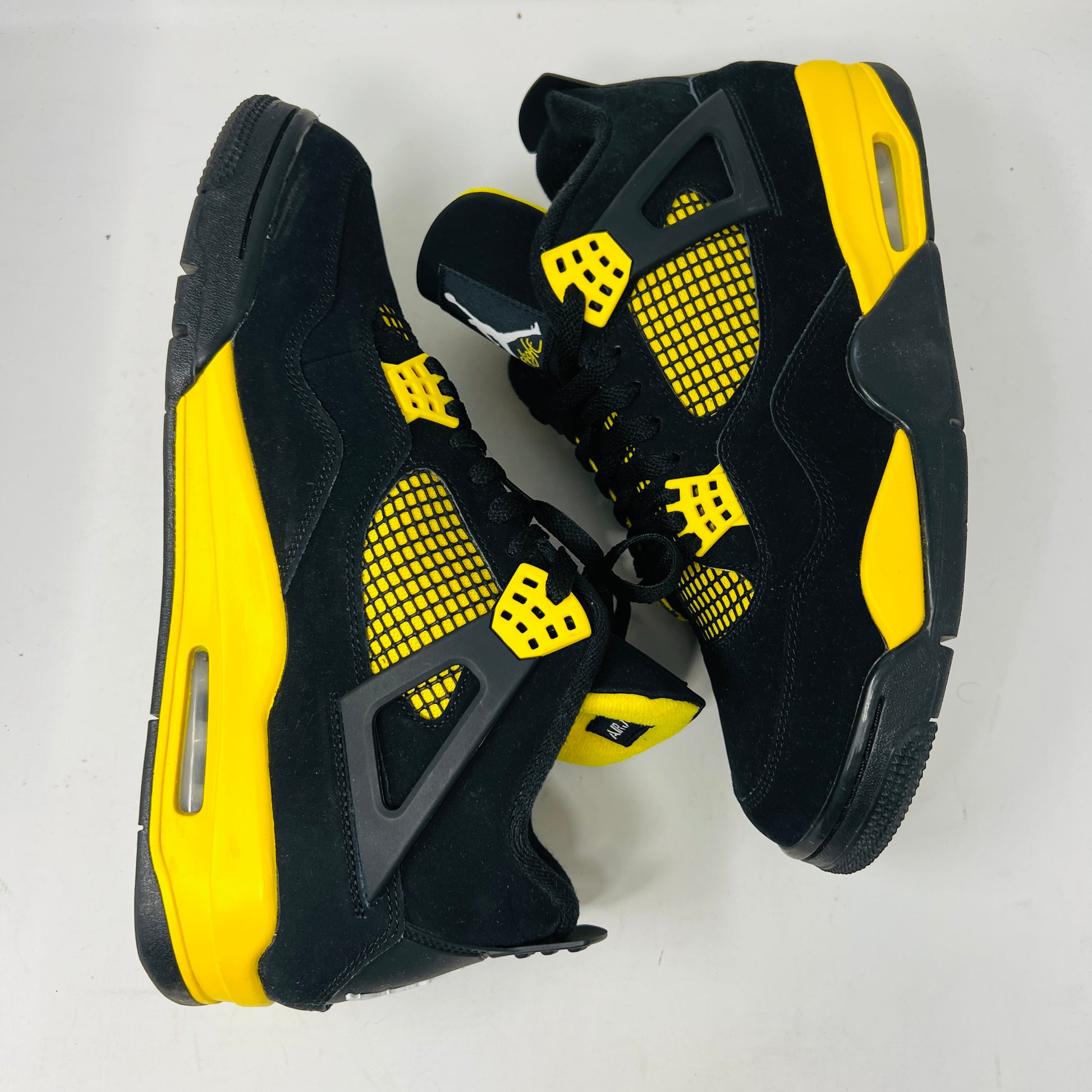 The Jordan 4 Retro Thunder (2023) features black uppers with yellow accents, a white sole, and visible air cushioning. It rests on a black and gray speckled shoebox in good condition and includes the distinctive wing design.