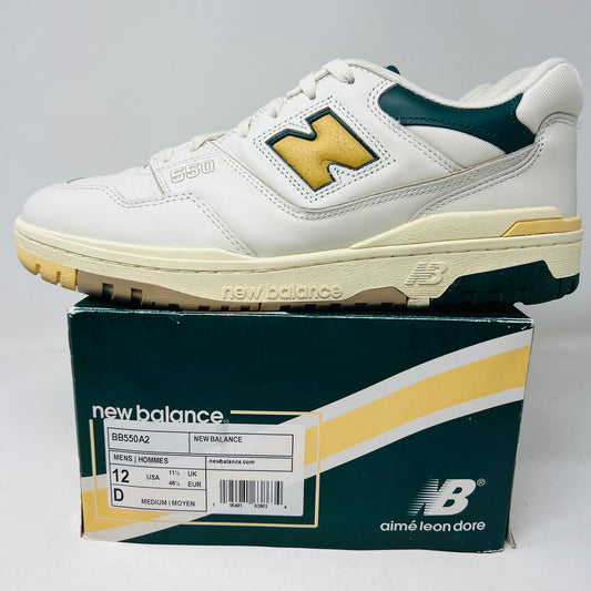 A pair of New Balance 550 Aime Leon Dore Natural Green sneakers in a white colorway with green and yellow accents sits on a matching shoebox. The sleek uppers add to their fresh aesthetic. They are size 12 and part of a collaboration with Aimé Leon Dore.