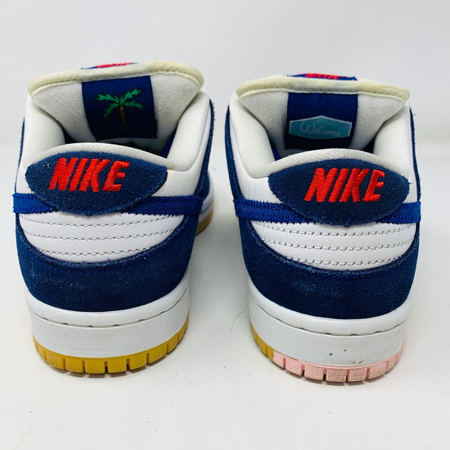 The image displays the rear view of Nike SB Dunk Low Los Angeles Dodgers sneakers by Nike, featuring a white and navy blue design. NIKE is embroidered in red on the heel tabs, and the soles vary: light brown on the left shoe and light pink on the right shoe.