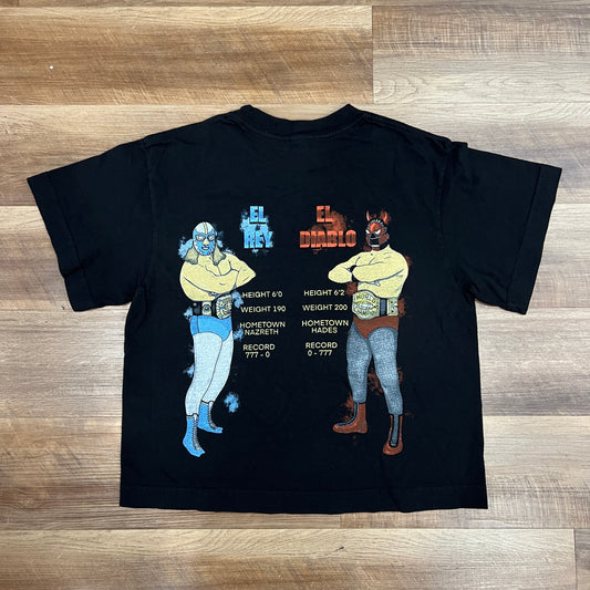 A Holy Ground black cropped Holy Rumble Tee, offered in sizes XS-XL, lies on a wooden floor. Bright yellow text on the left chest promotes The Big Cheese sporting event with captivating phrases like El Hombres.