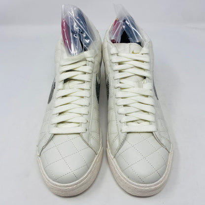 A pair of Nike SB Supreme Blazer White 2006 sneakers with quilted stitching, white laces, and a silver logo. Each shoe has a plastic-wrapped item inside, set against a plain white background.