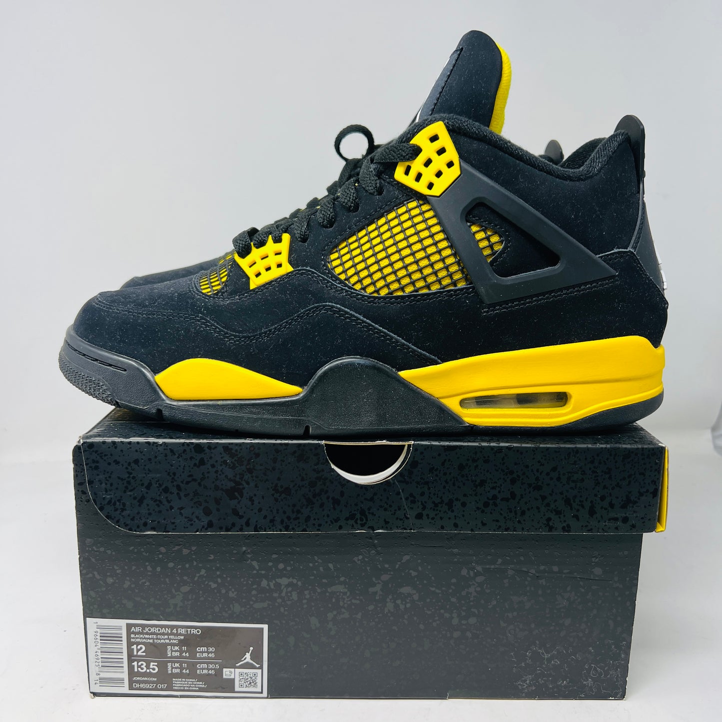 The Jordan 4 Retro Thunder (2023) features black uppers with yellow accents, a white sole, and visible air cushioning. It rests on a black and gray speckled shoebox in good condition and includes the distinctive wing design.