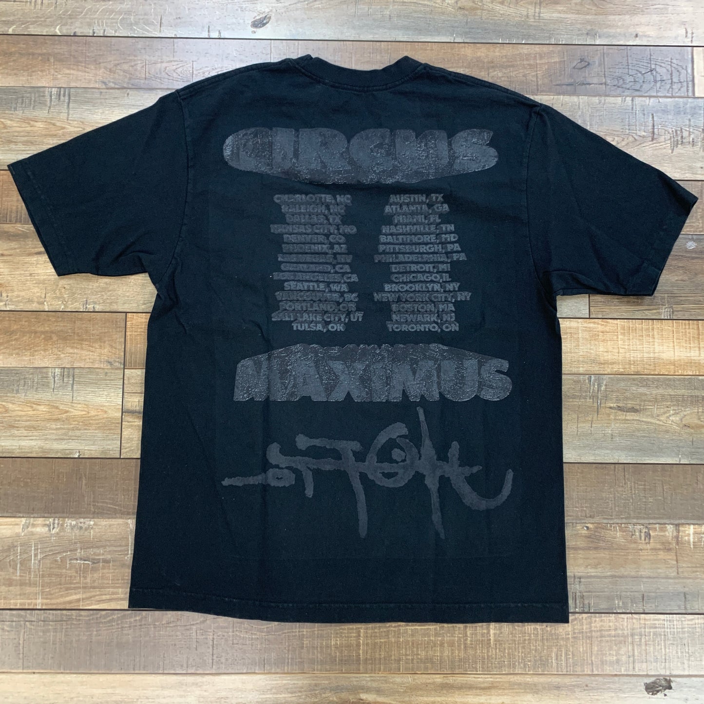 The Travis Scott Utopia Tour Tee by Cactus Jack, in black, is laid flat on a wooden floor. The back displays a list of cities in gray, framed by CIRCUS and MAXIMUS. A large gray signature-like design adorns the bottom.