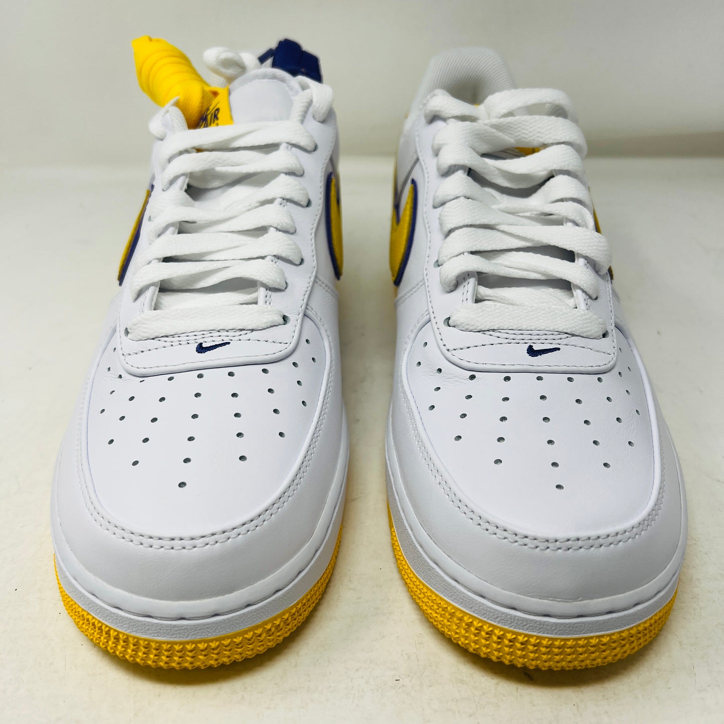 The Nike Air Force 1 Low Retro QS Kobe Bryant Lakers Home sneakers feature a white base with yellow accents, blue Nike logos, and perforated toe detailing, displayed side by side on a white background.