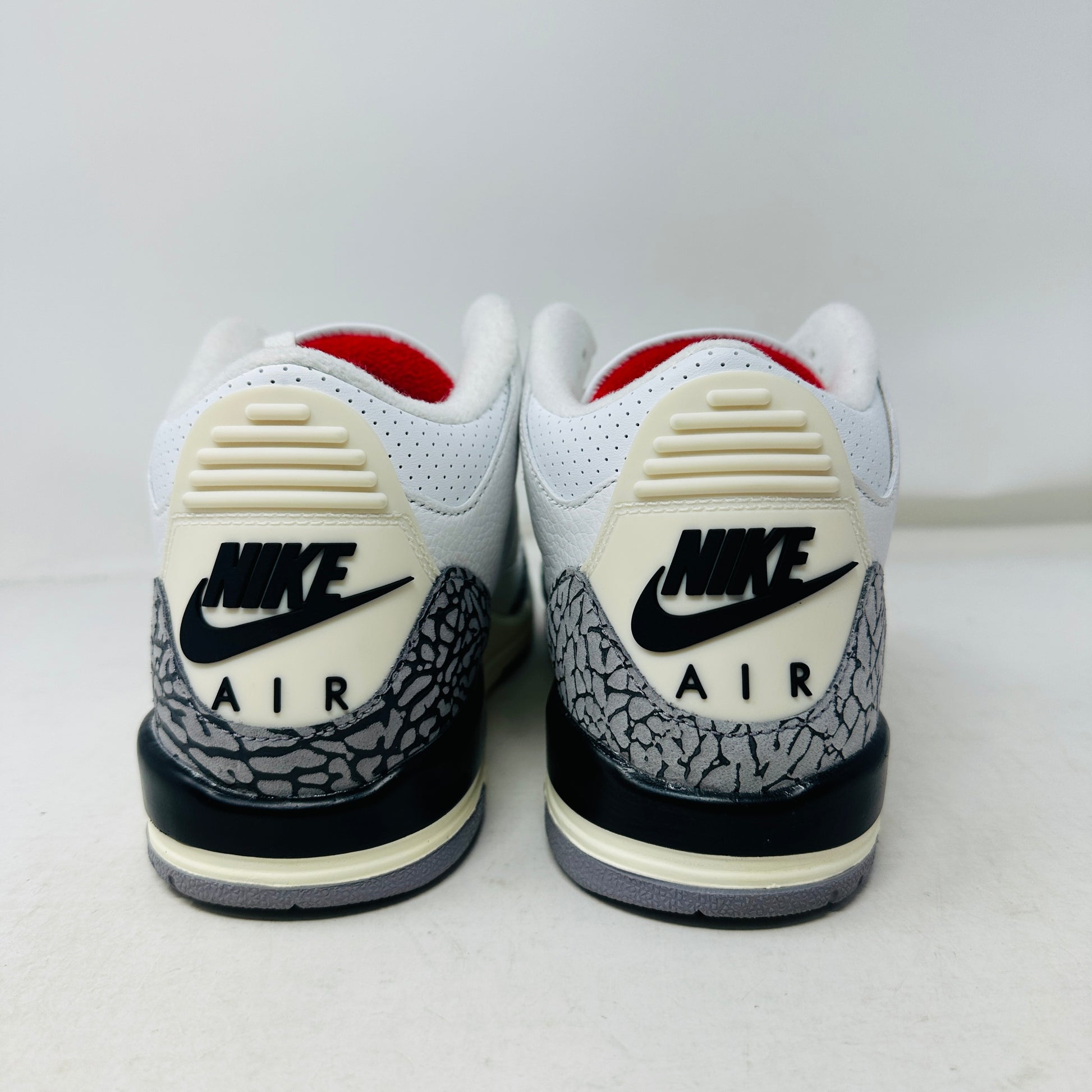 The image features the Jordan 3 Retro White Cement Reimagined (GS) sneakers. They showcase white leather uppers with gray cement patterns, black Nike Air logos, an off-white midsole, and red lining. The shoes are positioned on a white surface.