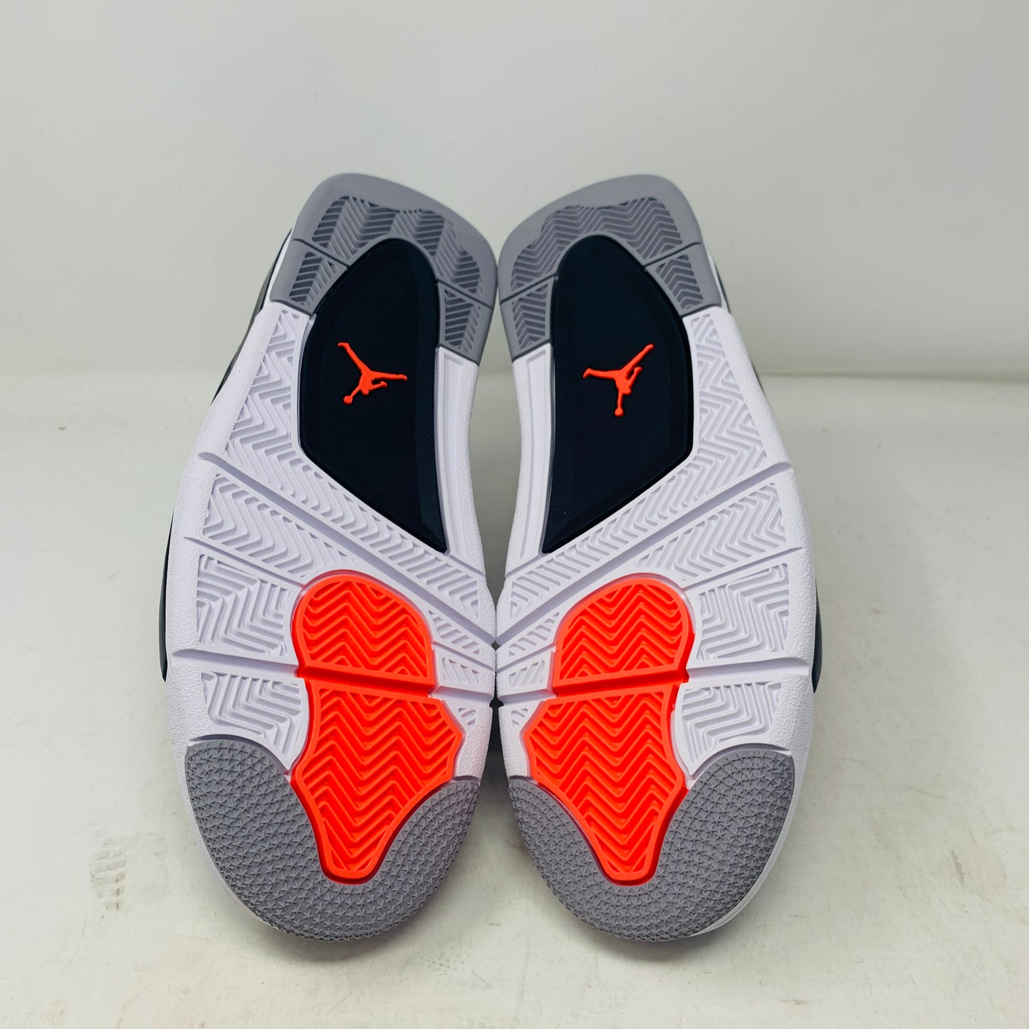 A pair of Jordan 4 Infrared sneakers by Jordan, featuring a gray mesh panel, black laces, and red accents with a distinctive logo on the tongue. They are positioned side by side on a white surface against a plain background.