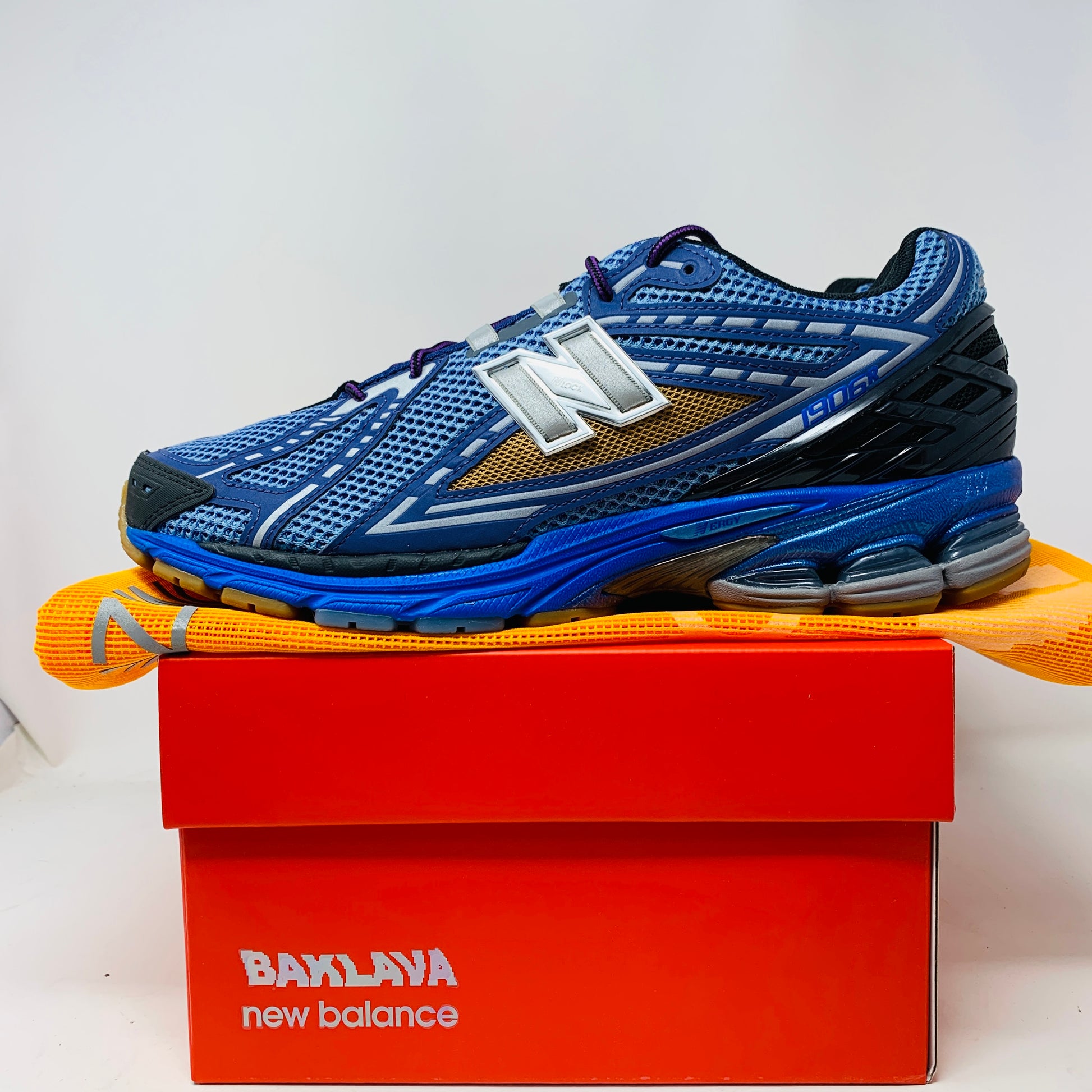 A brand new New Balance 1906R Action Bronson Medusa Azul sneaker with intricate designs and a silver N logo rests atop an orange box labeled BAKLAVA new balance. The 2024 edition features a layered design with black and metallic accents, complete with an intricate sole and dust bag.