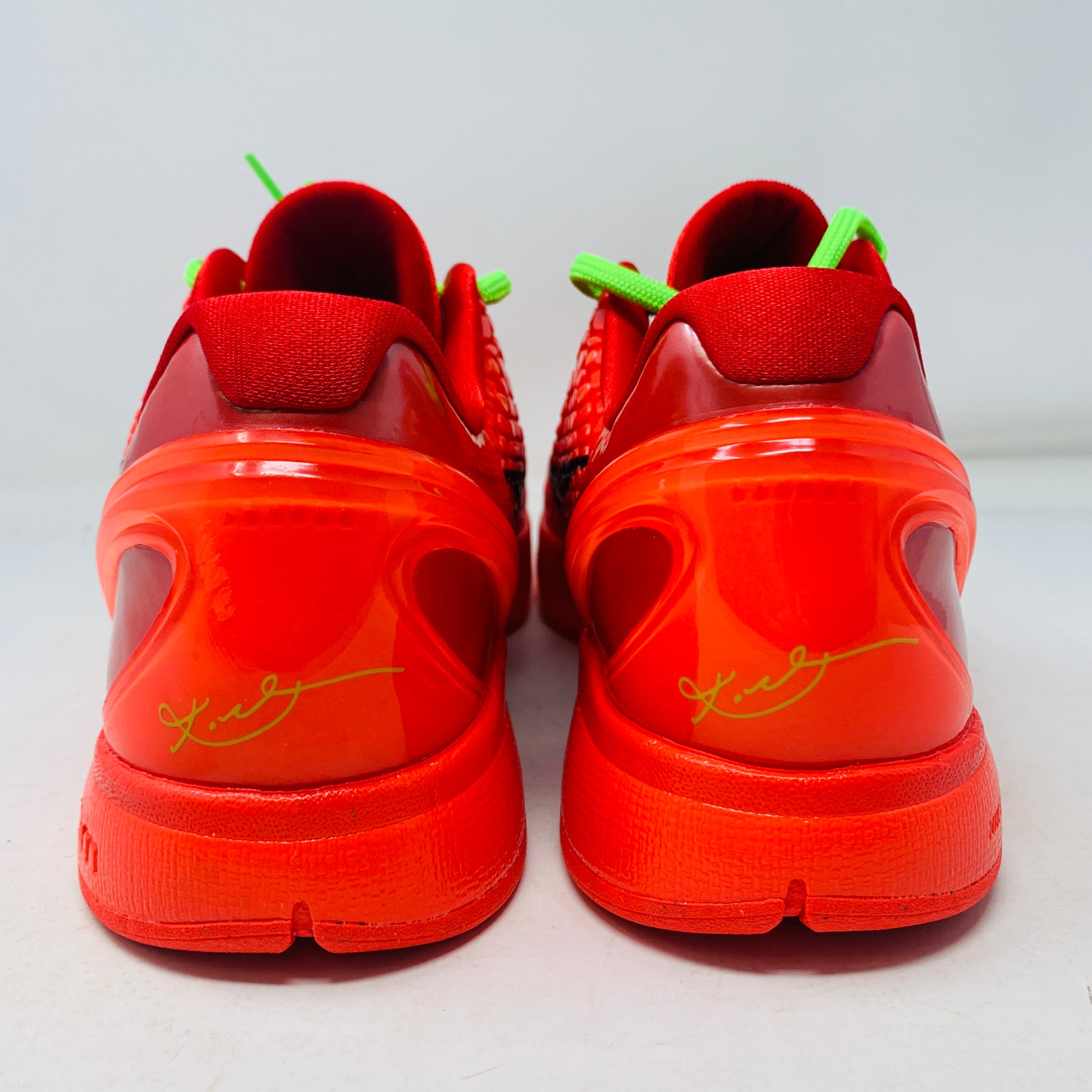 The Nike Kobe 6 Protro Reverse Grinch shoes, in bright red with neon green accents and signature details on the back, are displayed from the rear. Their textured design is set against a plain white surface.