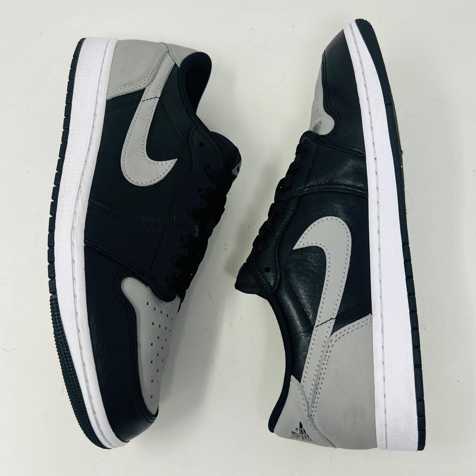 The Jordan 1 Retro Low OG Shadow (2024) sneakers in black and grey are displayed on a grey Nike box, showcasing clean uppers and matching laces. A small brown box complements the scene, with the visible size label enhancing authenticity.