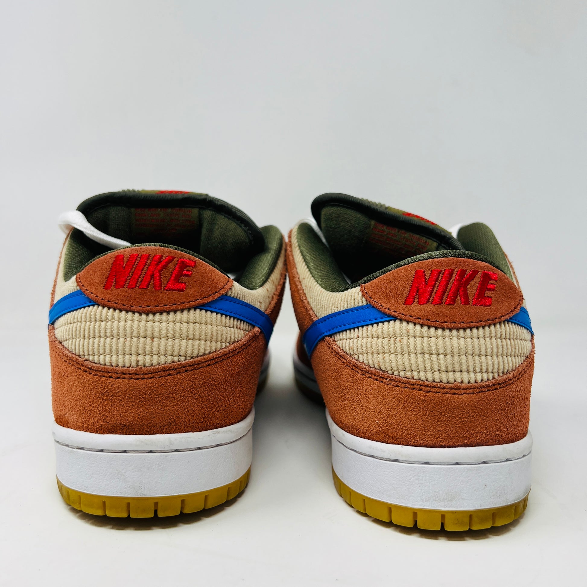 The Nike SB Dunk Low Corduroy Dusty Peach sneakers blend brown suede, beige corduroy, and blue accents with olive laces and white soles. Red NIKE text adorns the heel, above gum outsoles. Captured from the back against a plain backdrop, they embody clean lines and authentic style.