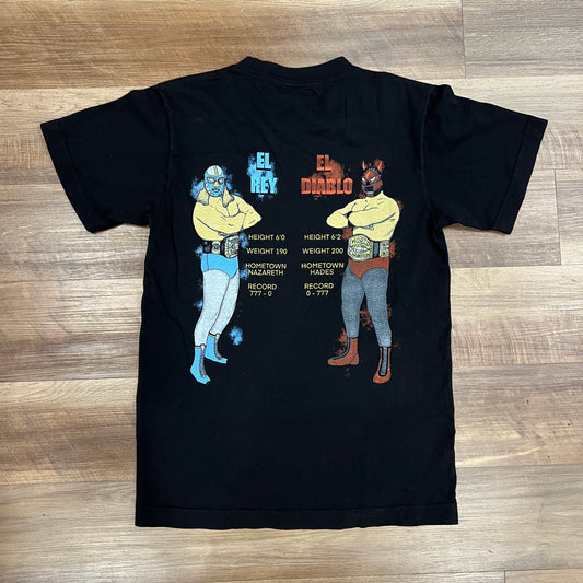 The Holy Rumble Tee by Holy Ground is a brand new black T-shirt, available in sizes S-XXL, featuring a yellow design with text and graphics on the left chest. Laid flat on a wooden floor.