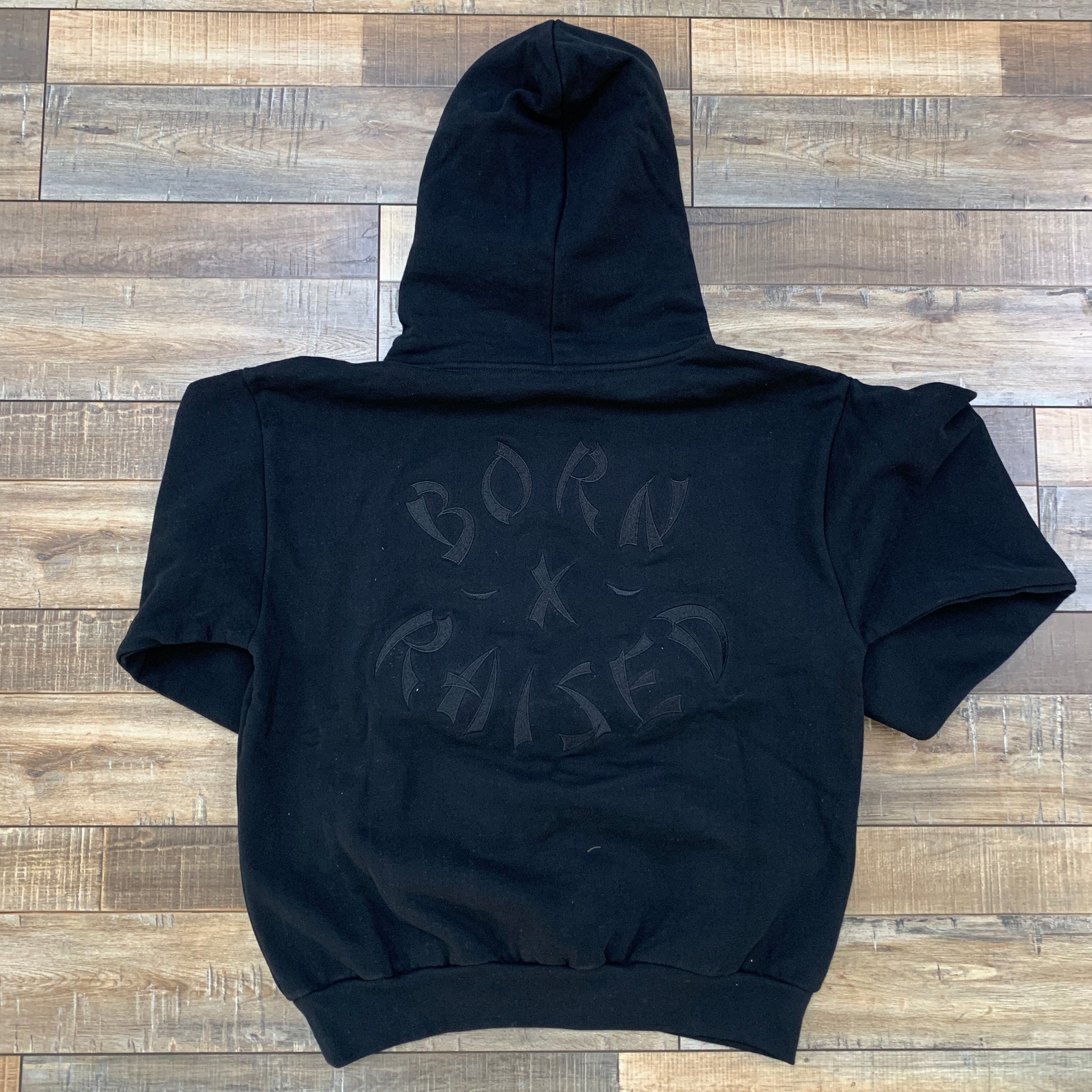 A Born x Raised Hoodie Black from born x raised is laid flat on a wooden floor, featuring BORN X RAISED printed on the back.