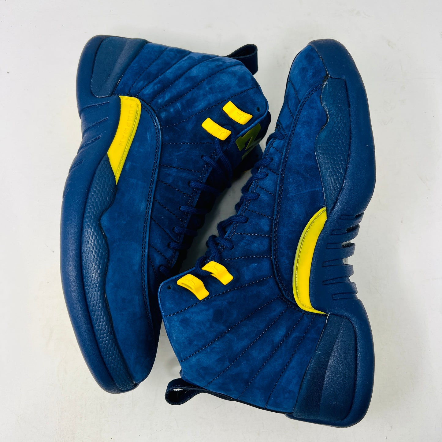 The Jordan 12 Retro Michigan is a blue high-top sneaker with yellow accents from Jordans 2018 Shoes collection. It features a textured surface, stitching details, and a visible Jumpman label. The dark blue sole highlights its vibrant design against a white background.