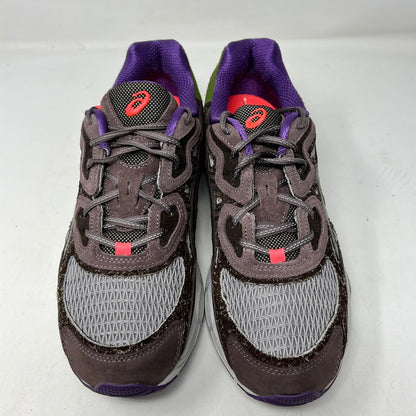 The ASICS Gel-NYC Bodega After-Hours sneakers, in brown with gray, purple, and green accents, sit on a box. A packet of extra red laces is attached. The size and details are prominently shown on the label.