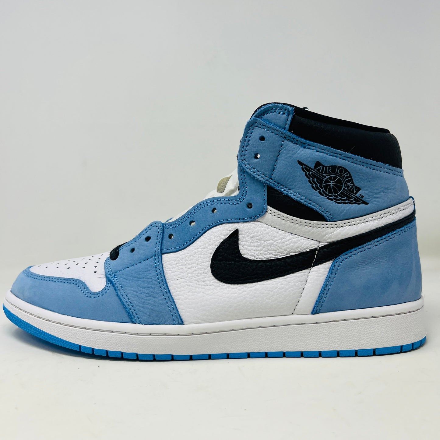 The side view of the Jordan 1 Retro High OG University Blue, by Jordan, showcases its blue, white, and black design. It features a prominent black Nike swoosh logo, blue laces, a blue sole, and a winged logo near the ankle on clean uppers against a plain white background.