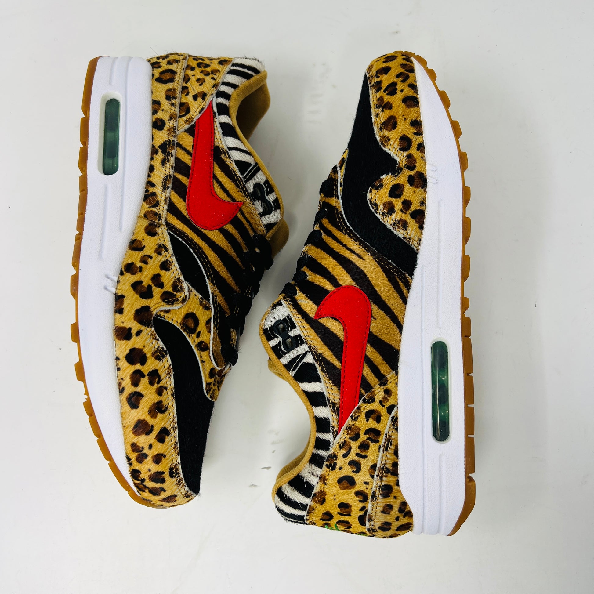 The Nike Air Max 1 Atmos Animal Pack 2.0 features leopard and zebra patterned sneakers with a vibrant orange swoosh, white sole with a transparent air unit, and brown outsole against a plain white background.