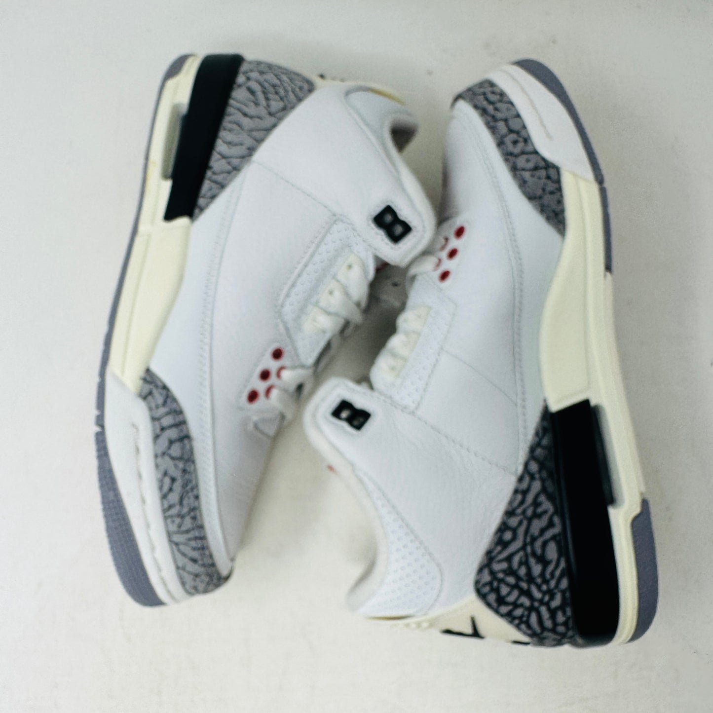 The Jordan 3 Retro White Cement Reimagined (GS) sneakers, by Jordan, are white with gray and black textured accents, red eyelets, and a black heel tab.