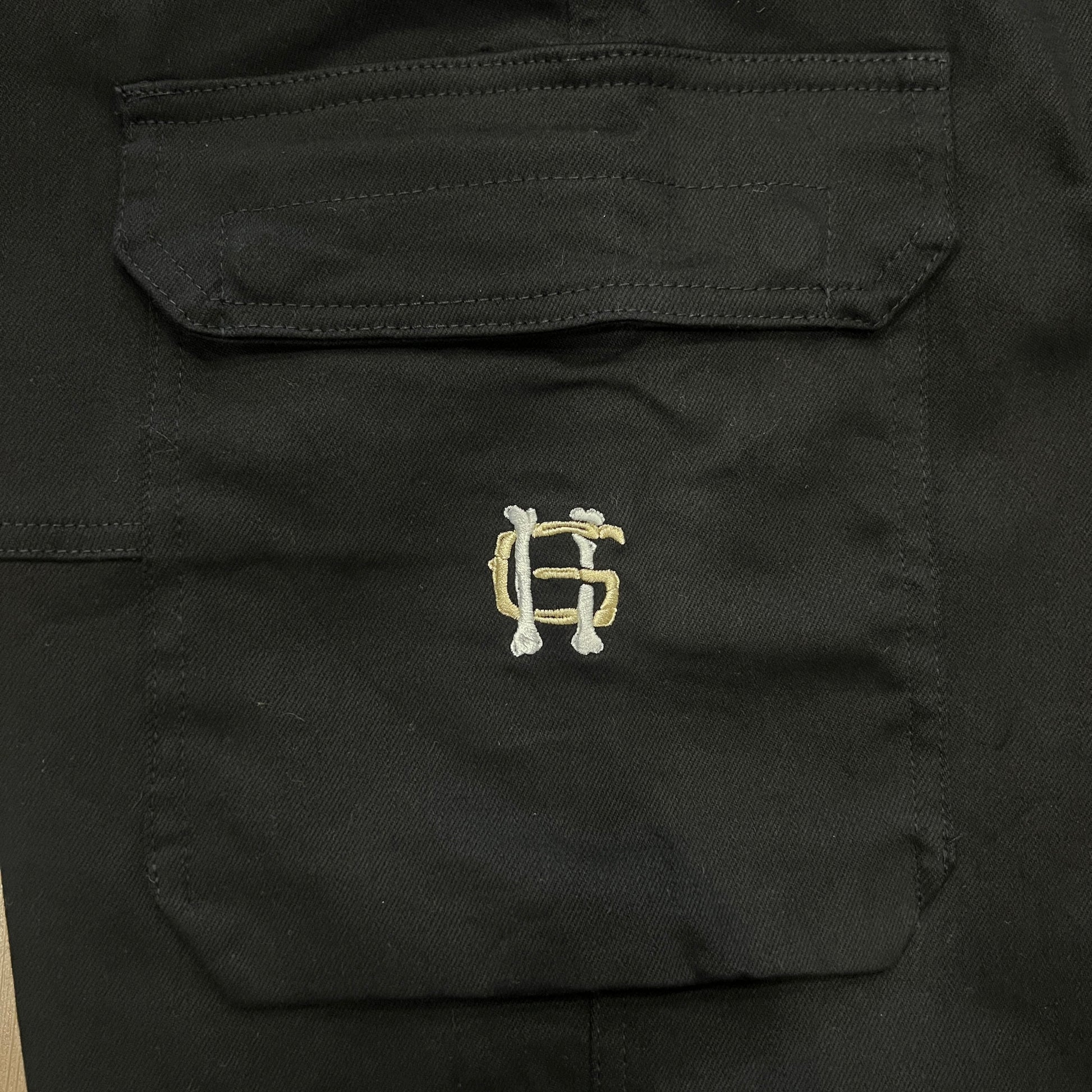 The Holy Ground Cargo Black pants, featuring large side pockets and a subtle shine, are on a wooden floor. Available in XS-XXL with a white logo on one pocket and a visible waist tag. Be sure to consult the size chart for the perfect fit.